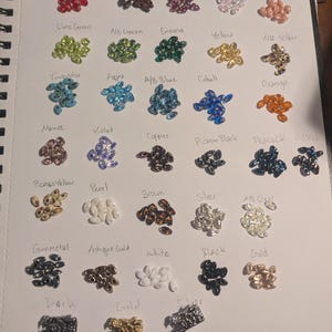 Op de afbeelding: Een pagina uit een notitieboek toont een verscheidenheid aan kleurrijke kralen, elk gelabeld met een andere kleurnaam. Kleuren zijn Baksteenrood, Limoengroen, Turkoois, Mauve, Picasso Geel, Gunmetal, Donker, Picasso Rood, A/B Groen, Aqua, Violet, Parel, Antiek Goud, Wit, Zwart, Goud, Fuchsia, Emerald, A/B Blauw, Koper, Bruin, Zilver, Roze, Geel, Kobalt, Picasso Zwart, A/B Helder, Zalm, A/B Geel, Oranje, Pauw en Olie.