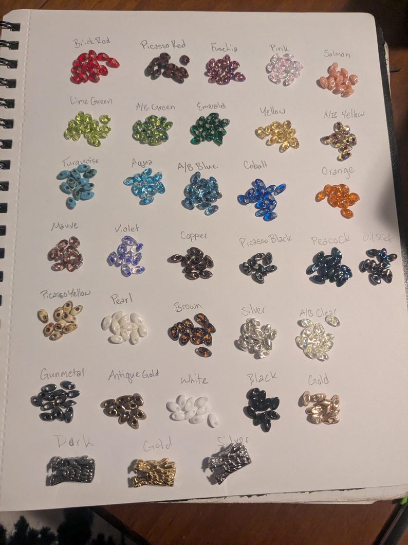 Op de afbeelding: Een pagina uit een notitieboek toont een verscheidenheid aan kleurrijke kralen, elk gelabeld met een andere kleurnaam. Kleuren zijn Baksteenrood, Limoengroen, Turkoois, Mauve, Picasso Geel, Gunmetal, Donker, Picasso Rood, A/B Groen, Aqua, Violet, Parel, Antiek Goud, Wit, Zwart, Goud, Fuchsia, Emerald, A/B Blauw, Koper, Bruin, Zilver, Roze, Geel, Kobalt, Picasso Zwart, A/B Helder, Zalm, A/B Geel, Oranje, Pauw en Olie.