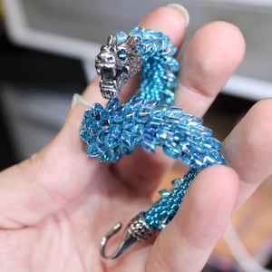 Op de afbeelding: Een kralen drakenarmband in blauw- en zilvertinten. De kop en sluiting van de draak zijn zilverkleurig, terwijl het lichaam is gemaakt van iriserende blauwe kralen. De armband is ongeveer 18 cm lang.