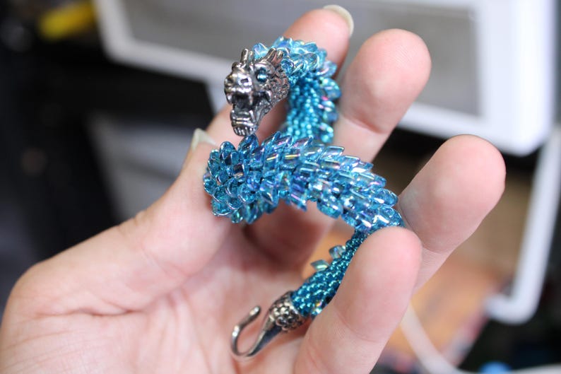Op de afbeelding: Een kralen drakenarmband in blauw- en zilvertinten. De kop en sluiting van de draak zijn zilverkleurig, terwijl het lichaam is gemaakt van iriserende blauwe kralen. De armband is ongeveer 18 cm lang.