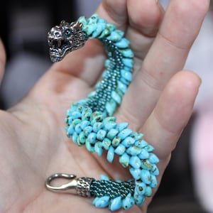 May include: A turquoise beaded bracelet in the shape of a dragon. The dragon's head is silver with a detailed design. The bracelet is held in a hand, showcasing the intricate beadwork and the silver clasp.