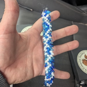 Op de afbeelding: Een armband met blauwe en witte kralen, vastgehouden in een hand. De kralen zijn gerangschikt in een herhalend patroon, waardoor een textuur ontstaat. Het ontwerp van de armband is ingewikkeld, met een contrast tussen de blauwe en witte kralen. Ongeveer 18 cm lang.