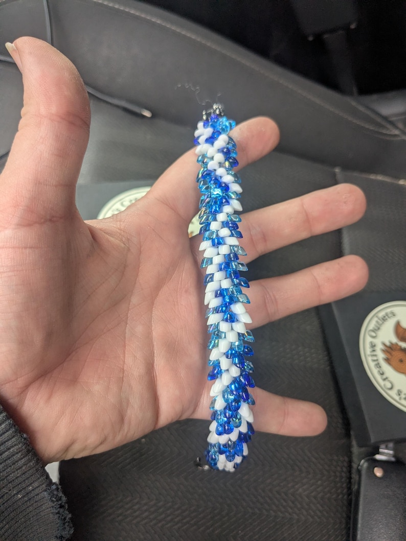 Op de afbeelding: Een armband met blauwe en witte kralen, vastgehouden in een hand. De kralen zijn gerangschikt in een herhalend patroon, waardoor een textuur ontstaat. Het ontwerp van de armband is ingewikkeld, met een contrast tussen de blauwe en witte kralen. Ongeveer 18 cm lang.