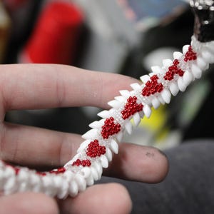 Op de afbeelding: Een handgemaakte armband met een witte basis en rode kralenaccenten. De armband heeft een uniek ontwerp met een reeks witte, tandachtige vormen, en de rode kralen zijn gerangschikt in hartvormige patronen. De sluiting is zilver.