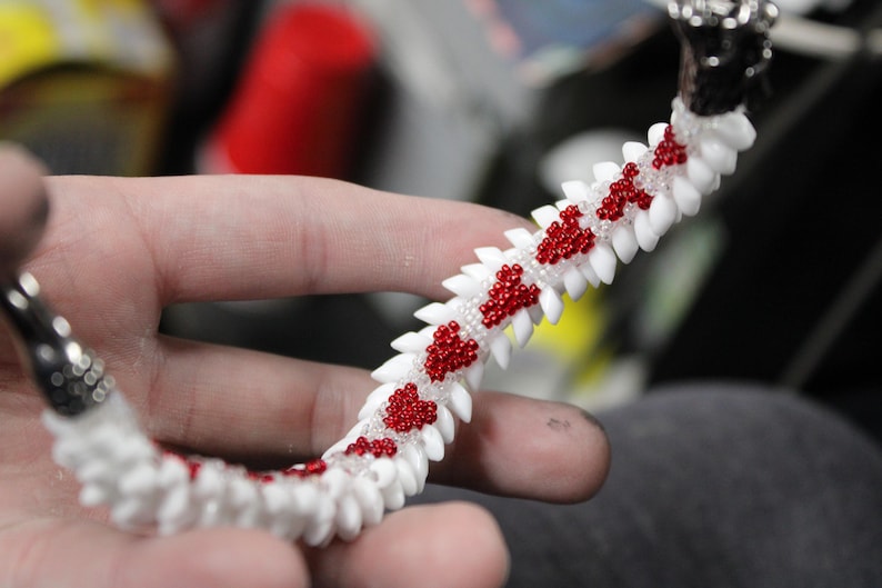 Op de afbeelding: Een handgemaakte armband met een witte basis en rode kralenaccenten. De armband heeft een uniek ontwerp met een reeks witte, tandachtige vormen, en de rode kralen zijn gerangschikt in hartvormige patronen. De sluiting is zilver.