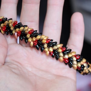 Op de afbeelding: Een armband met zwarte, rode en gouden kralen. De kralen zijn in een herhalend patroon gerangschikt. De armband wordt in een hand gehouden, waarbij het ingewikkelde ontwerp en de kleurencombinatie worden getoond. De kralen cre&euml;ren een textuureffect.