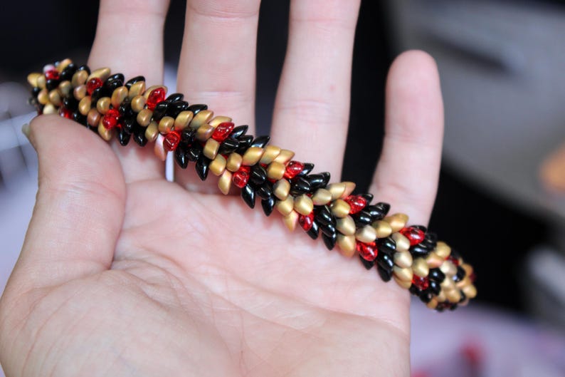 Op de afbeelding: Een armband met zwarte, rode en gouden kralen. De kralen zijn in een herhalend patroon gerangschikt. De armband wordt in een hand gehouden, waarbij het ingewikkelde ontwerp en de kleurencombinatie worden getoond. De kralen cre&euml;ren een textuureffect.