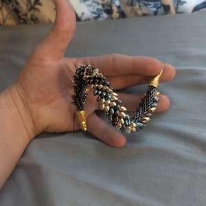 Op de afbeelding: Een kralenarmband met een gouden sluiting en accenten. De armband heeft een patroon van zwarte, gouden en blauwe kralen. De armband is ongeveer 18 cm lang.