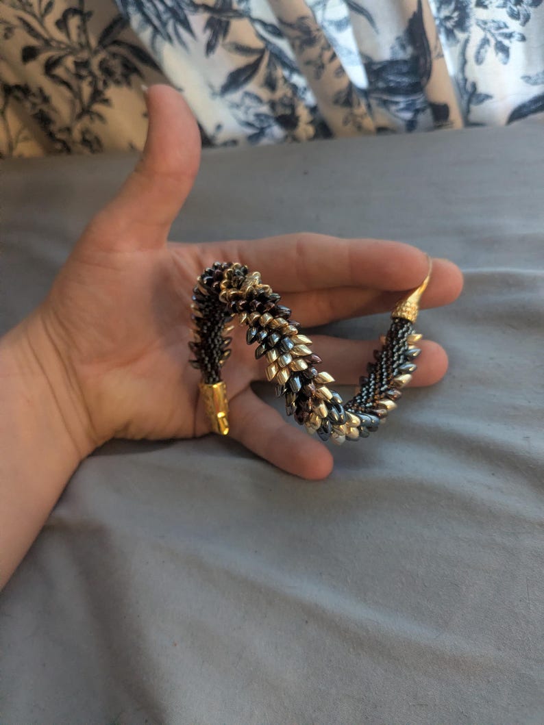 Op de afbeelding: Een kralenarmband met een gouden sluiting en accenten. De armband heeft een patroon van zwarte, gouden en blauwe kralen. De armband is ongeveer 18 cm lang.