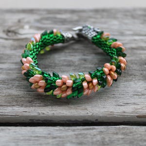 Op de afbeelding: Een kralenarmband met een geweven ontwerp. Het heeft groene, perzik en transparante kralen en een zilverkleurige sluiting. De kralen zijn gerangschikt in een bloemmotief. De armband ligt op een verweerd houten oppervlak.