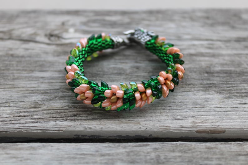 Op de afbeelding: Een kralenarmband met een geweven ontwerp. Het heeft groene, perzik en transparante kralen en een zilverkleurige sluiting. De kralen zijn gerangschikt in een bloemmotief. De armband ligt op een verweerd houten oppervlak.