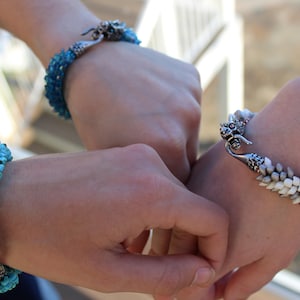 Op de afbeelding: Close-up van drie armbanden. E&eacute;n armband is turquoise, een andere is wit en grijs, en de derde is blauw. Elke armband heeft een uniek ontwerp en sluiting. De armbanden zitten om polsen.
