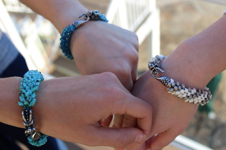 Op de afbeelding: Close-up van drie armbanden. E&eacute;n armband is turquoise, een andere is wit en grijs, en de derde is blauw. Elke armband heeft een uniek ontwerp en sluiting. De armbanden zitten om polsen.