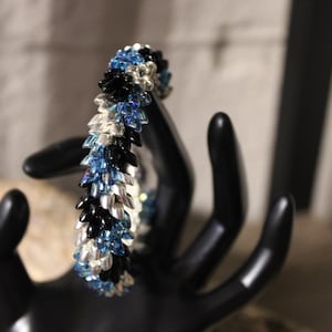 Op de afbeelding: Een kralenarmband met een bladachtig ontwerp. De armband heeft een combinatie van zwarte, zilveren en blauwe kralen. De armband wordt gepresenteerd op een zwarte handvormige sieradenhouder.