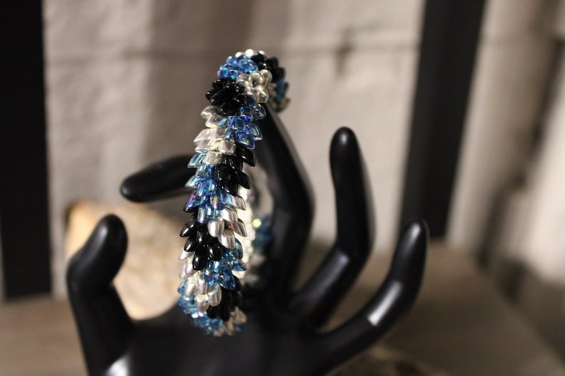 Op de afbeelding: Een kralenarmband met een bladachtig ontwerp. De armband heeft een combinatie van zwarte, zilveren en blauwe kralen. De armband wordt gepresenteerd op een zwarte handvormige sieradenhouder.