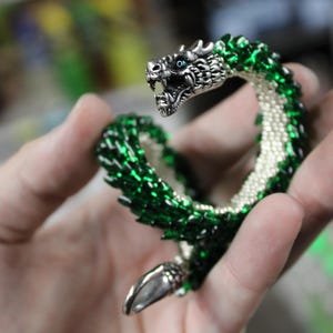 Op de afbeelding: Een gedetailleerde drakenarmband met een zilveren kop en staart. De body is gemaakt van groene kralen met een witte kralenbuik. De kop van de draak heeft blauwe ogen en een open mond.