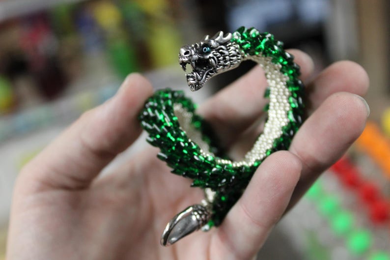 Op de afbeelding: Een gedetailleerde drakenarmband met een zilveren kop en staart. De body is gemaakt van groene kralen met een witte kralenbuik. De kop van de draak heeft blauwe ogen en een open mond.