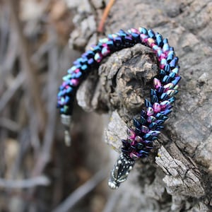 Op de afbeelding: Een kralenarmband met een iriserende afwerking in blauwe, paarse en roze tinten. De armband is gedrapeerd over een getextureerde, grijsbruine tak. De sluiting is zilverkleurig.