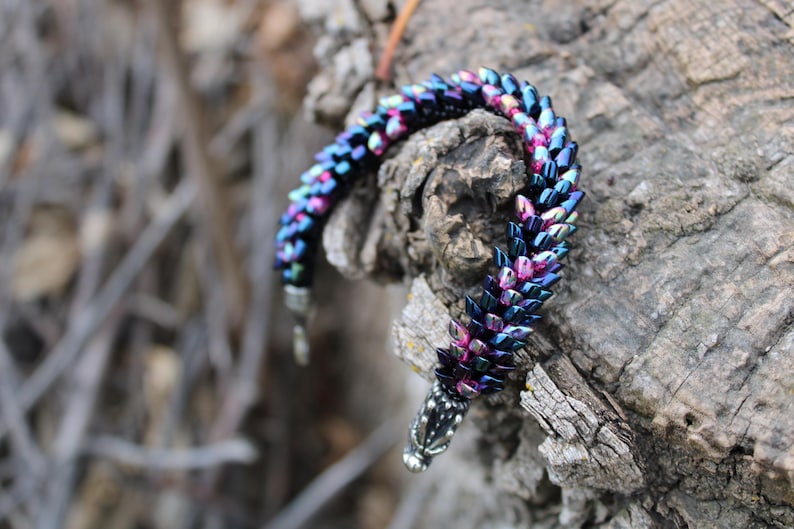 Op de afbeelding: Een kralenarmband met een iriserende afwerking in blauwe, paarse en roze tinten. De armband is gedrapeerd over een getextureerde, grijsbruine tak. De sluiting is zilverkleurig.