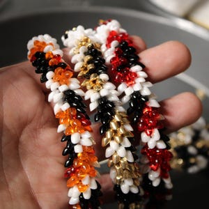 Op de afbeelding: Close-up van verschillende kralenarmbanden in verschillende kleuren. E&eacute;n armband heeft zwarte, oranje en witte kralen. Een andere heeft zwarte, gouden en witte kralen. De derde armband is rood, wit en zwart. De armbanden worden in een hand gehouden.