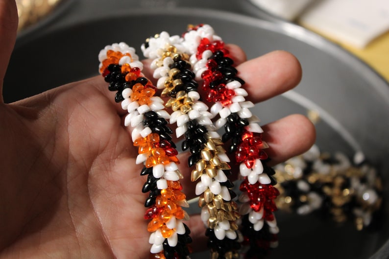 Op de afbeelding: Close-up van verschillende kralenarmbanden in verschillende kleuren. E&eacute;n armband heeft zwarte, oranje en witte kralen. Een andere heeft zwarte, gouden en witte kralen. De derde armband is rood, wit en zwart. De armbanden worden in een hand gehouden.