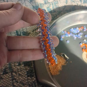 Op de afbeelding: Een kralen knutselproject in uitvoering. Het stuk is een lange, smalle strook met afwisselende rijen oranje en blauwe kralen. Een naald en draad zijn aan de bovenkant bevestigd. Een metalen bakje bevat losse kralen in oranje en blauw.