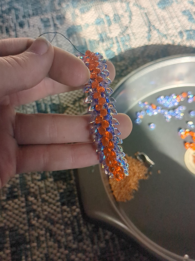 Op de afbeelding: Een kralen knutselproject in uitvoering. Het stuk is een lange, smalle strook met afwisselende rijen oranje en blauwe kralen. Een naald en draad zijn aan de bovenkant bevestigd. Een metalen bakje bevat losse kralen in oranje en blauw.