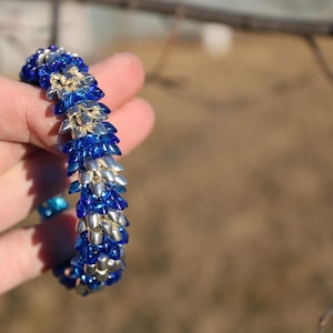 Op de afbeelding: Een armband van blauwe en zilveren kralen, vastgehouden in een hand. De kralen zijn gerangschikt in een herhalend patroon. De achtergrond is wazig. De kralen van de armband cre&euml;ren een textuur.