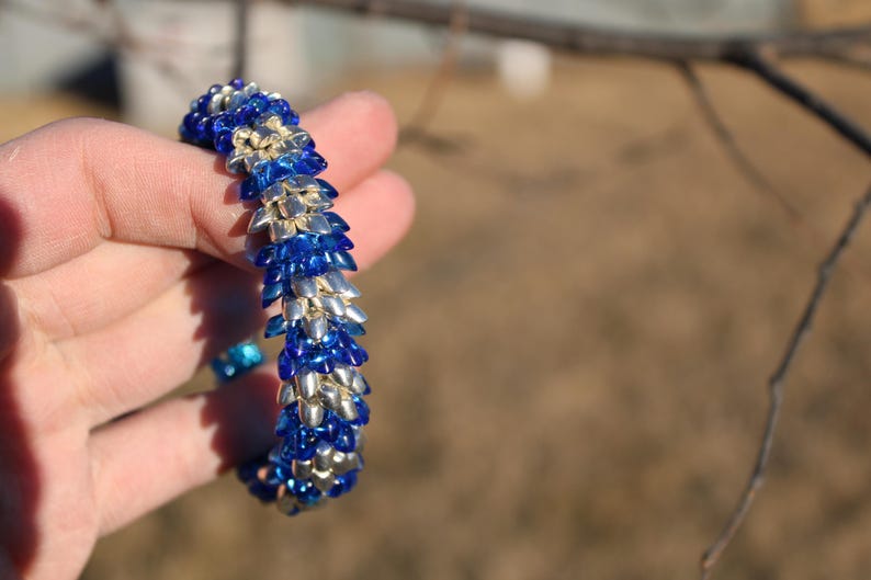 Op de afbeelding: Een armband van blauwe en zilveren kralen, vastgehouden in een hand. De kralen zijn gerangschikt in een herhalend patroon. De achtergrond is wazig. De kralen van de armband cre&euml;ren een textuur.