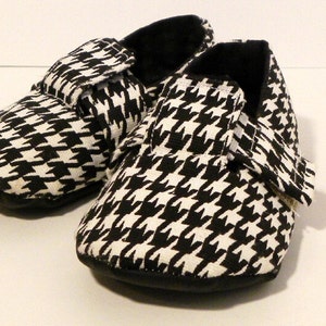 Puede incluir: Un par de zapatos de bebé en tejido negro y blanco con estampado de pata de gallo y cierre de velcro.