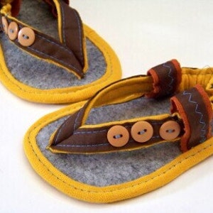 May include: A pair of brown felt baby sandals with yellow trim and brown buttons. The sandals have a strap that goes around the ankle and a toe post.