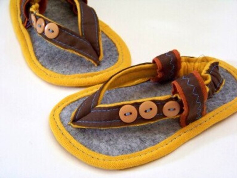 May include: A pair of brown felt baby sandals with yellow trim and brown buttons. The sandals have a strap that goes around the ankle and a toe post.