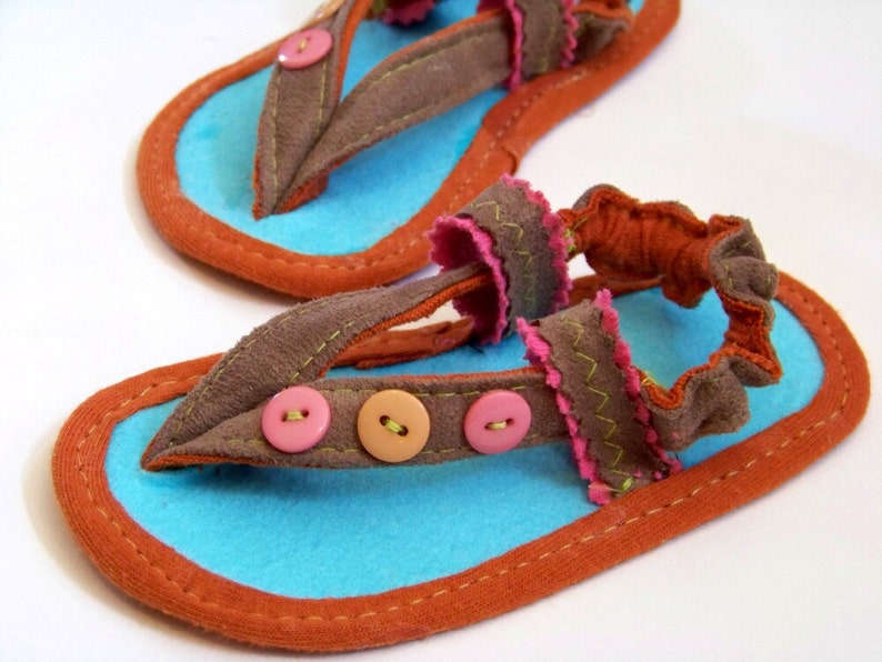 May include: A pair of brown suede sandals with blue and orange trim. The sandals have a decorative ruffle and three buttons on the strap.