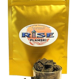 May include: A gold resealable bag of RISE PLAMSKI Shrimp Farm ornamental shrimp food. The bag has a label that says "100% ORGANIC and Natural Ingredients". A small glass bowl filled with dark green shrimp food flakes sits on a white surface.