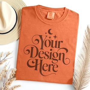 May include: An orange t-shirt with the text "Your Design Here" in a brown, decorative font. The shirt is next to a straw hat and dried plants. The shirt has a tag that says XL-70-80.