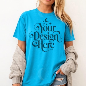 May include: A turquoise t-shirt with the text "Your Design Here" in a dark blue, decorative font. The shirt is short-sleeved and has a crew neck. A light gray cardigan is draped over the shoulders.