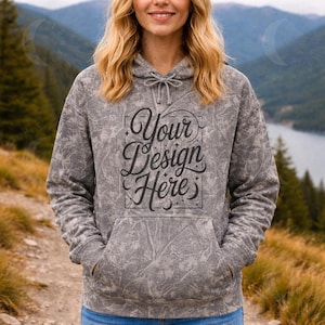 Puede incluir: Una sudadera con capucha gris con un patrón texturizado y un bolsillo delantero. La sudadera con capucha presenta el texto "Your Design Here" en una escritura decorativa. El modelo lleva vaqueros azules. El fondo muestra una montaña y un lago.