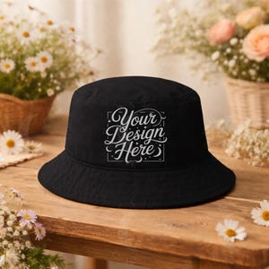 May include: Black bucket hat with the text "Your Design Here" in a decorative font. The hat is sitting on a wooden surface, with flowers in the background. The hat is a classic style, suitable for various occasions.