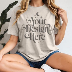 以下が含まれることがあります： 装飾的なグレーのフォントで「Your Design Here」というテキストが入った、明るいベージュのTシャツ。Tシャツは半袖とクルーネックです。モデルは青いデニムショーツと白いスニーカーを着用しています。