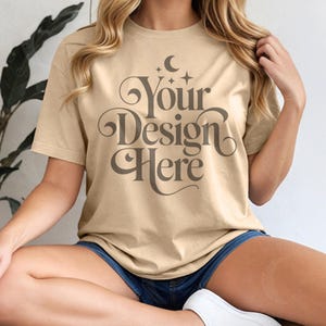 May include: Tan-colored t-shirt with the text "Your Design Here" in a decorative font. The shirt has short sleeves and a crew neck. The model is wearing blue denim shorts.