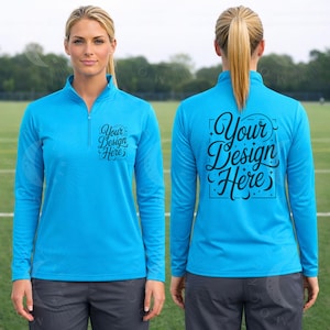 Puede incluir: Una camiseta deportiva turquesa de manga larga con cuello con cremallera de un cuarto. La parte delantera y trasera de la camiseta presentan el texto "Your Design Here" en una fuente decorativa. La modelo lleva pantalones deportivos grises.