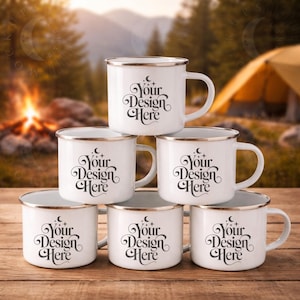 May include: A stack of six white enamel camping mugs with gold rims and handles. Each mug features the text "Your Design Here" in a decorative font. The mugs are arranged on a wooden surface with a blurred outdoor background.