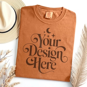 May include: A rust-colored t-shirt with the text "Your Design Here" in a decorative font. The shirt is laid flat on a white surface, with a straw hat and dried plants in the background. The shirt has a tag that reads XL-TG-EG.
