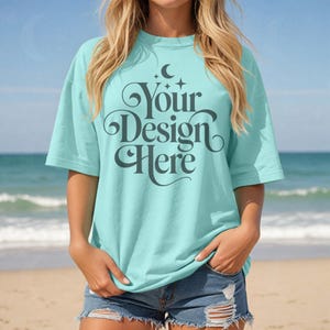May include: A light blue oversized t-shirt with the text "Your Design Here" in a decorative font. The shirt is being worn by a person on a beach.