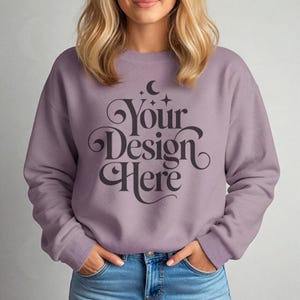 Puede incluir: Una sudadera de color malva con el texto "Your Design Here" en una escritura decorativa. La sudadera de manga larga tiene cuello redondo. La modelo lleva vaqueros azules.