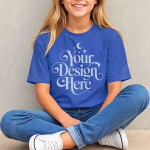 May include: A royal blue t-shirt with the text "Your Design Here" in a white, decorative font. The person is wearing blue jeans and white sneakers. The person is sitting with their legs crossed.