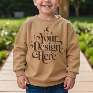 May include: A tan-colored sweatshirt with the text "Your Design Here" in a decorative font. The sweatshirt has long sleeves and a crew neck. The child is wearing blue jeans.