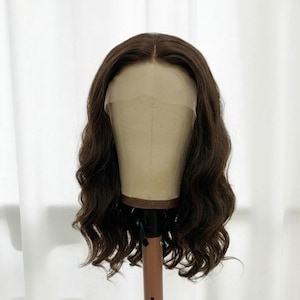 May include: A shoulder-length brown wig with a center part and loose, wavy texture. The wig features a sheer lace front panel that extends across the forehead, designed to mimic a natural hairline, displayed on a beige mannequin head.