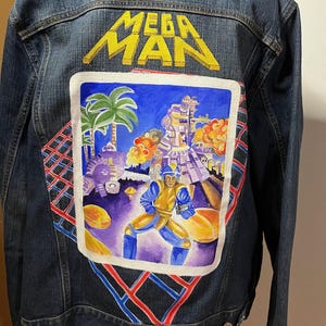 Puede incluir: Una chaqueta vaquera azul con un diseño pintado a mano del videojuego Mega Man. El diseño presenta una ciudad pixelada con una palmera, un robot y Mega Man en primer plano. Las palabras "MEGA MAN" están pintadas en amarillo en la parte posterior de la chaqueta.