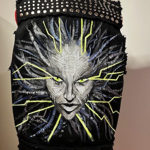 Puede incluir: Un chaleco vaquero negro con tachuelas plateadas y un diseño pintado de una cara con líneas amarillas brillantes y acentos azules y morados.